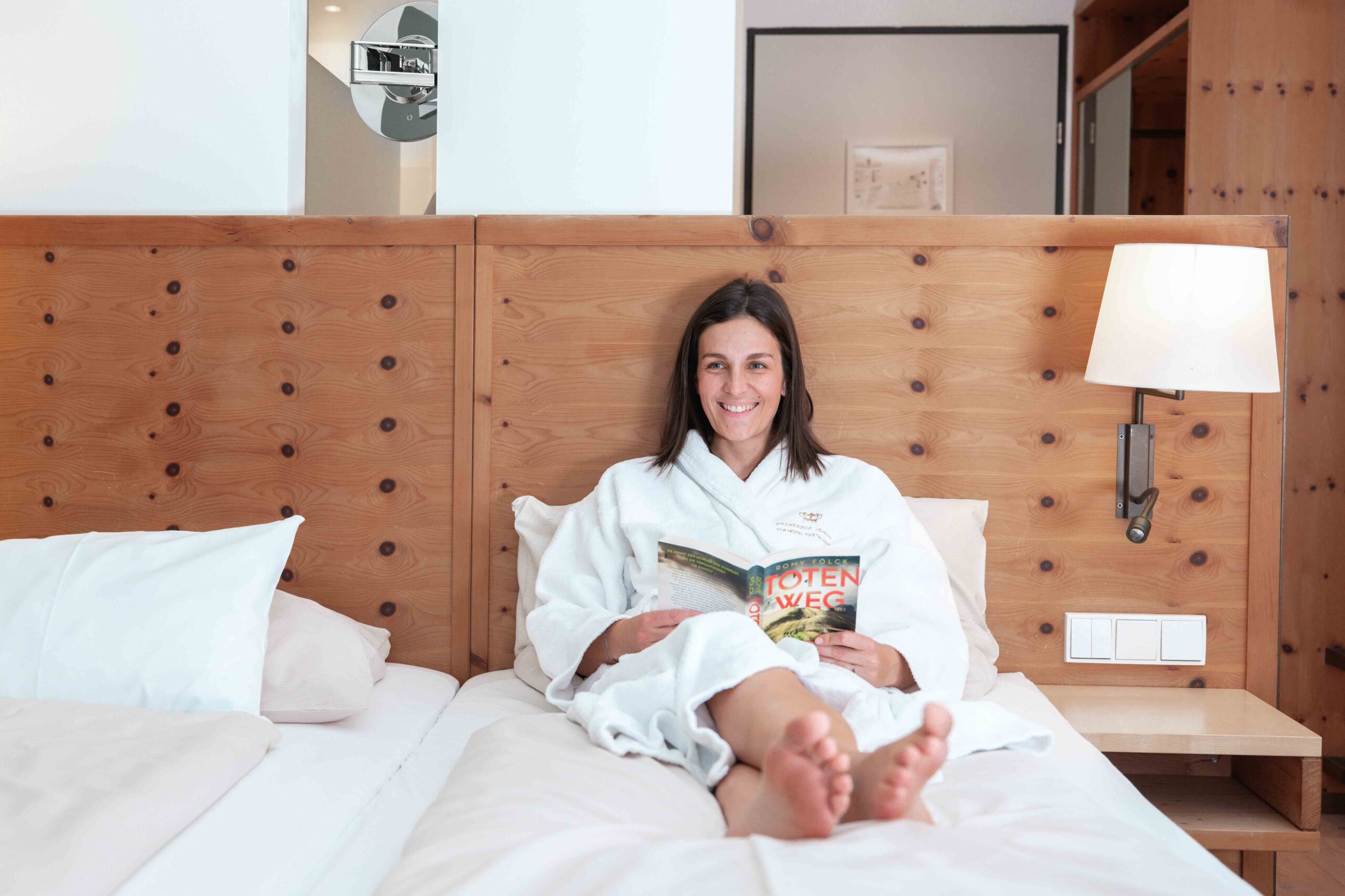 Eine Frau im Bademantel liegt gemütlich im Zirbenbett mit einem Buch.